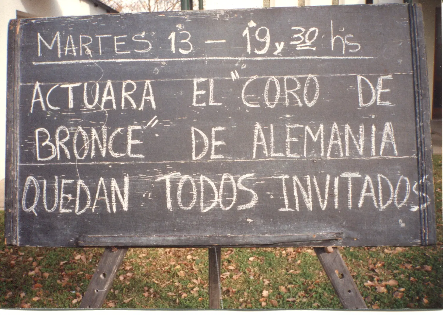 Pizarrón de madera sobre caballetes, escrito con tiza: Martes 13, 19,30 hs, actuará el Coro de Bronce de Alemania, quedan todos invitados
