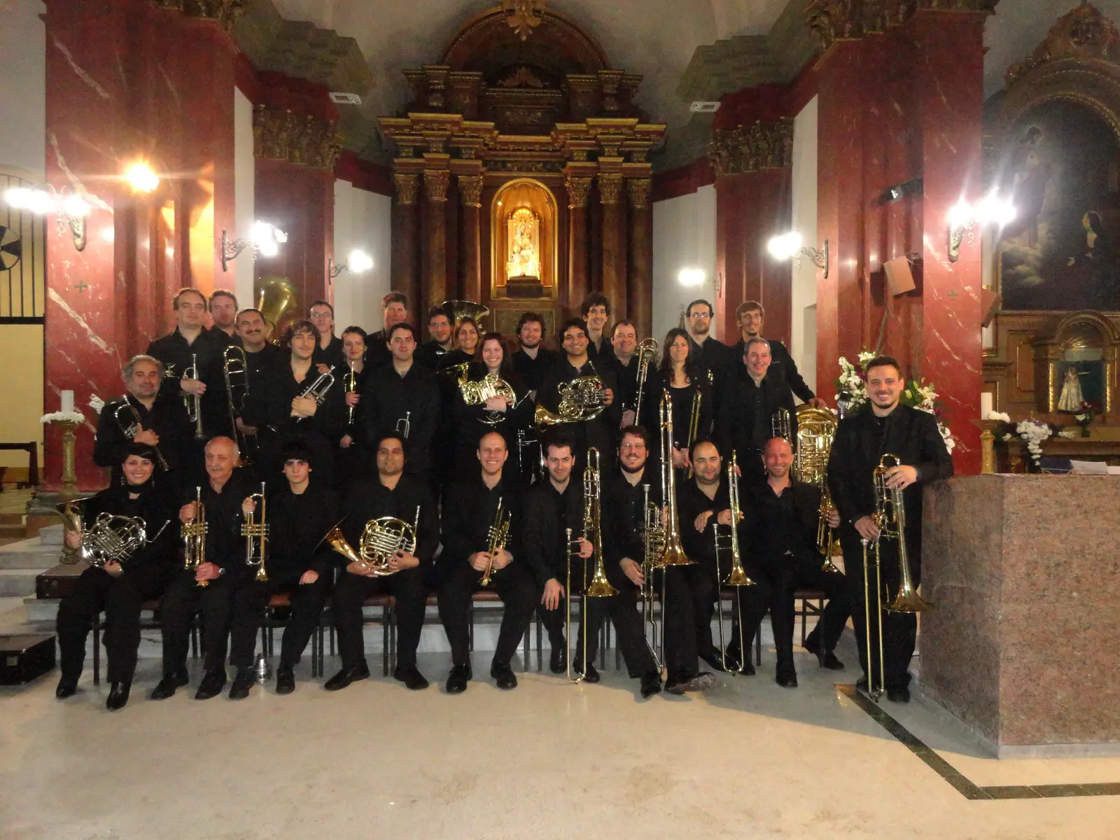 Unos treinta y cinco músicos vestidos de negro posando en tres filas en el presbiterio de una iglesia de mármol rojo y altar barroco dorado, todos con su instrumento