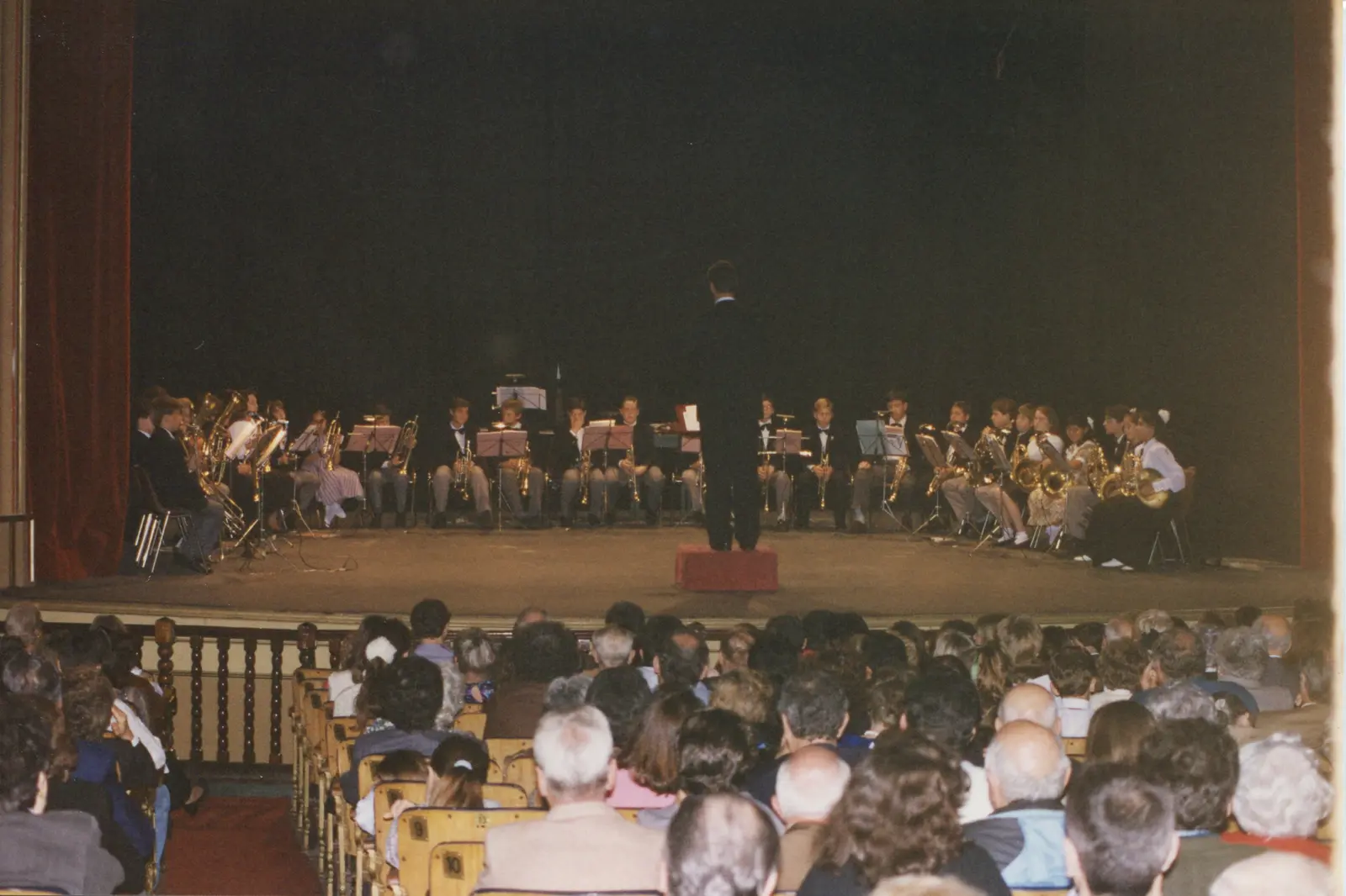 Vista desde la platea llena hacia el escenario del Teatro Coliseo, con unos veinticinco chicos sentados tocando y el director sobre una tarima roja