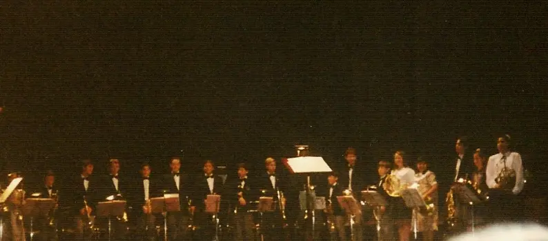 Fotografía apaisada y oscura de una veintena de músicos sentados en semicírculo sobre el escenario de un teatro, de ropa oscura, con los atriles iluminados y la sala a oscuras