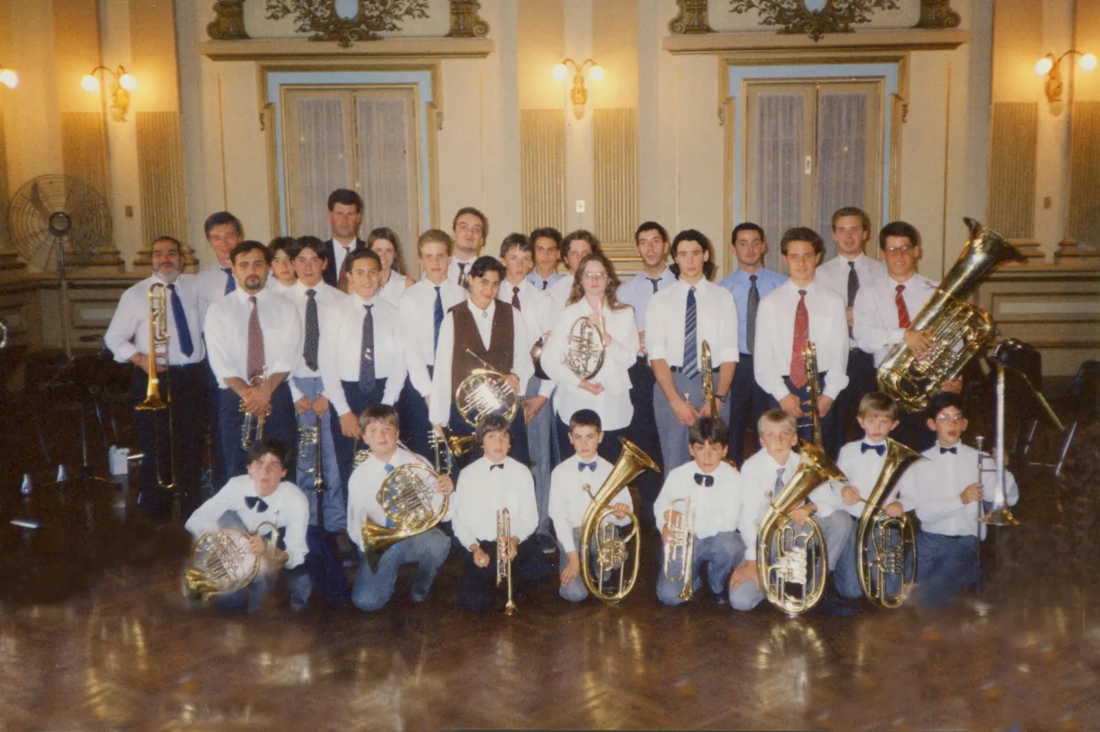 Fotografía en color de unos veintiocho músicos de bronces posando en un salón dorado de techo alto, con apliques de pared, molduras doradas y piso de parquet; ocho chicos arrodillados adelante con sus instrumentos