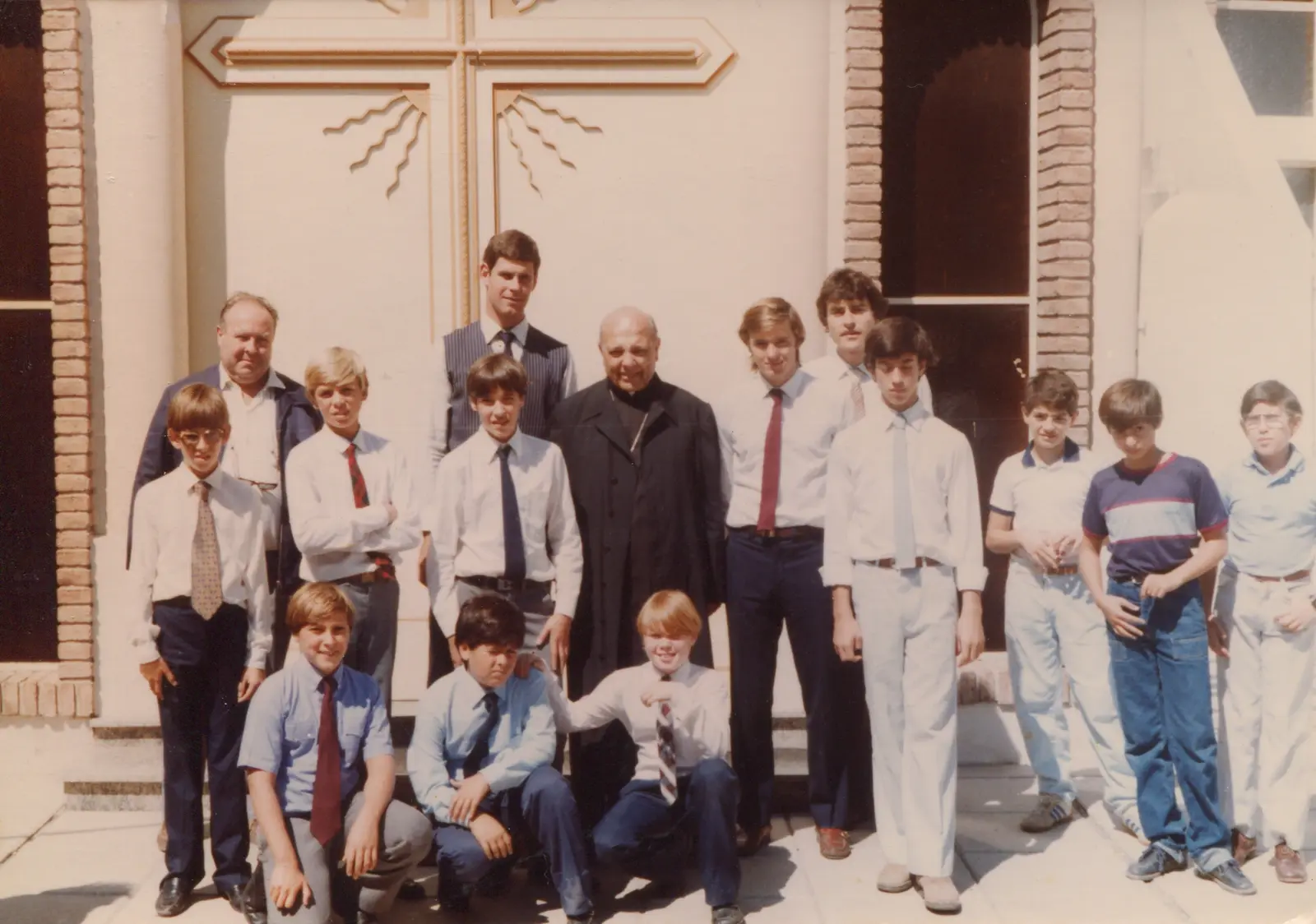Fotografía en color de dieciséis músicos, en su mayoría chicos, posando al sol frente a las puertas de una iglesia con una gran cruz dorada con rayos, junto a un clérigo mayor de sotana negra