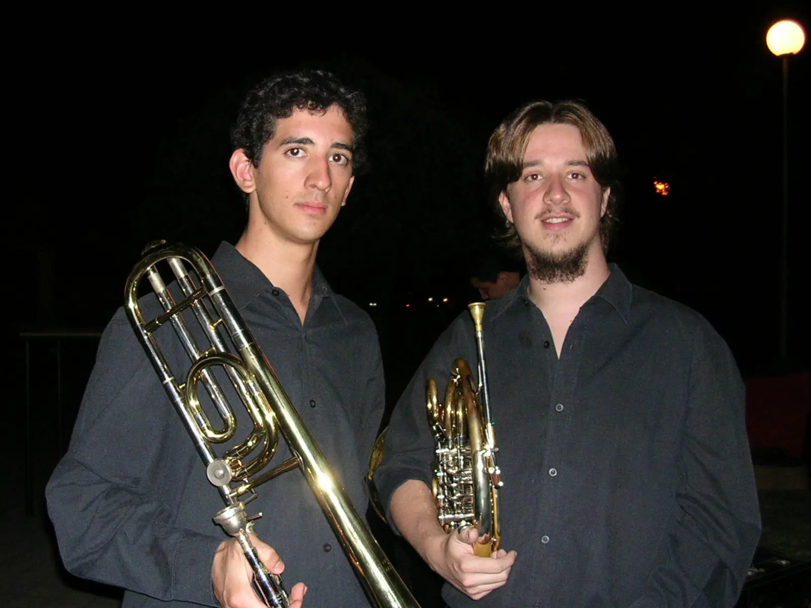 Fotografía nocturna de dos músicos jóvenes de camisa gris oscura al aire libre, uno con un trombón de varas y el otro con un fliscorno, bajo la luz anaranjada de una farola