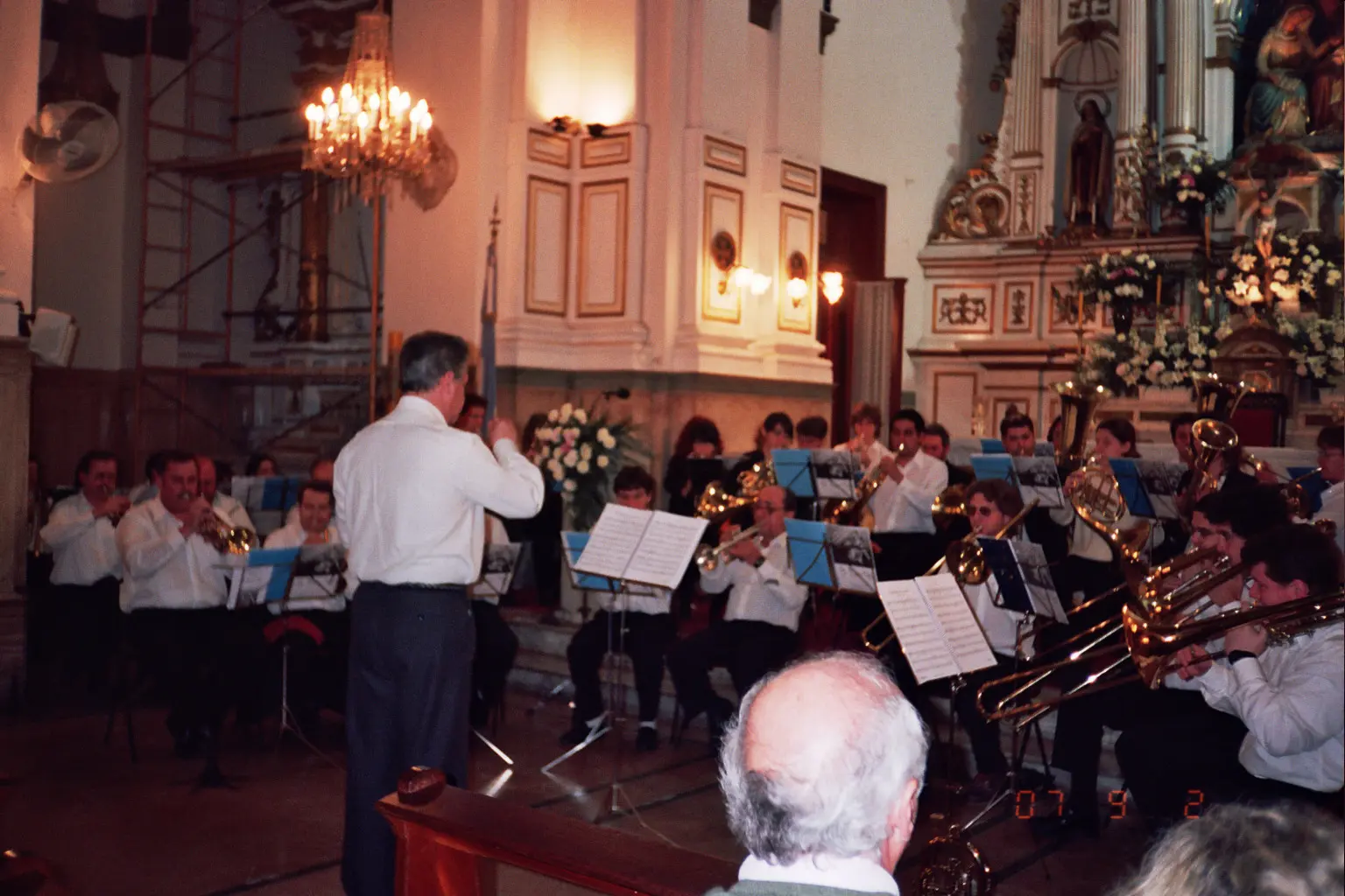 Unos treinta músicos de camisa blanca tocando dentro de una iglesia barroca con araña de caño, bandera argentina y altar dorado con flores, el director de espaldas en el centro