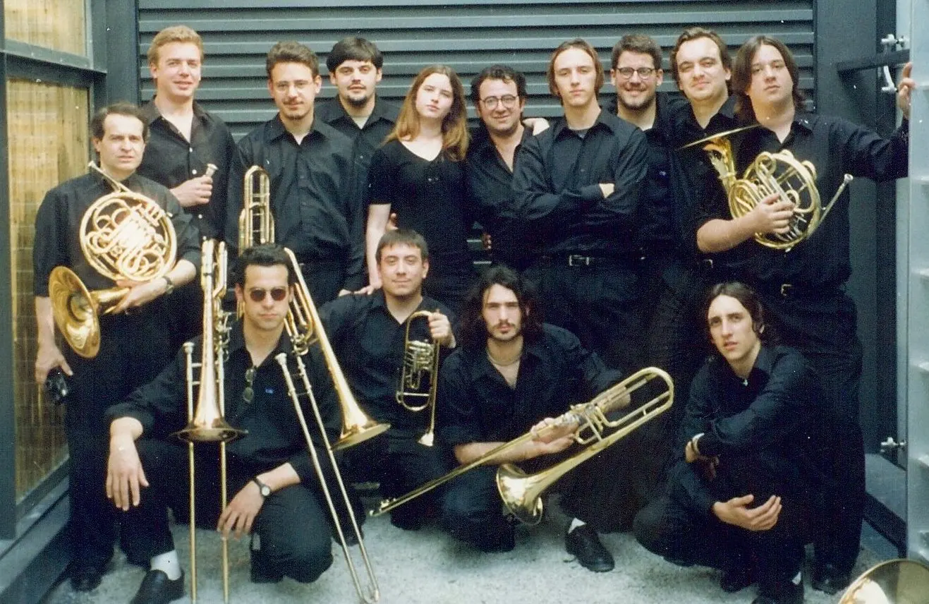 Quince músicos vestidos íntegramente de negro posando con sus instrumentos —dos cornos, trombones, trompetas y un fliscorno— contra una pared moderna de listones metálicos grises