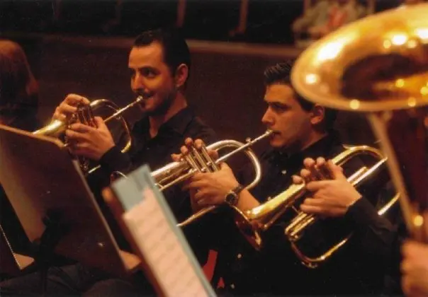 Primer plano de dos trompetistas de negro tocando bajo luz cálida de escenario, con el pabellón de una tuba en primer plano y una partitura de renglones azules en el atril