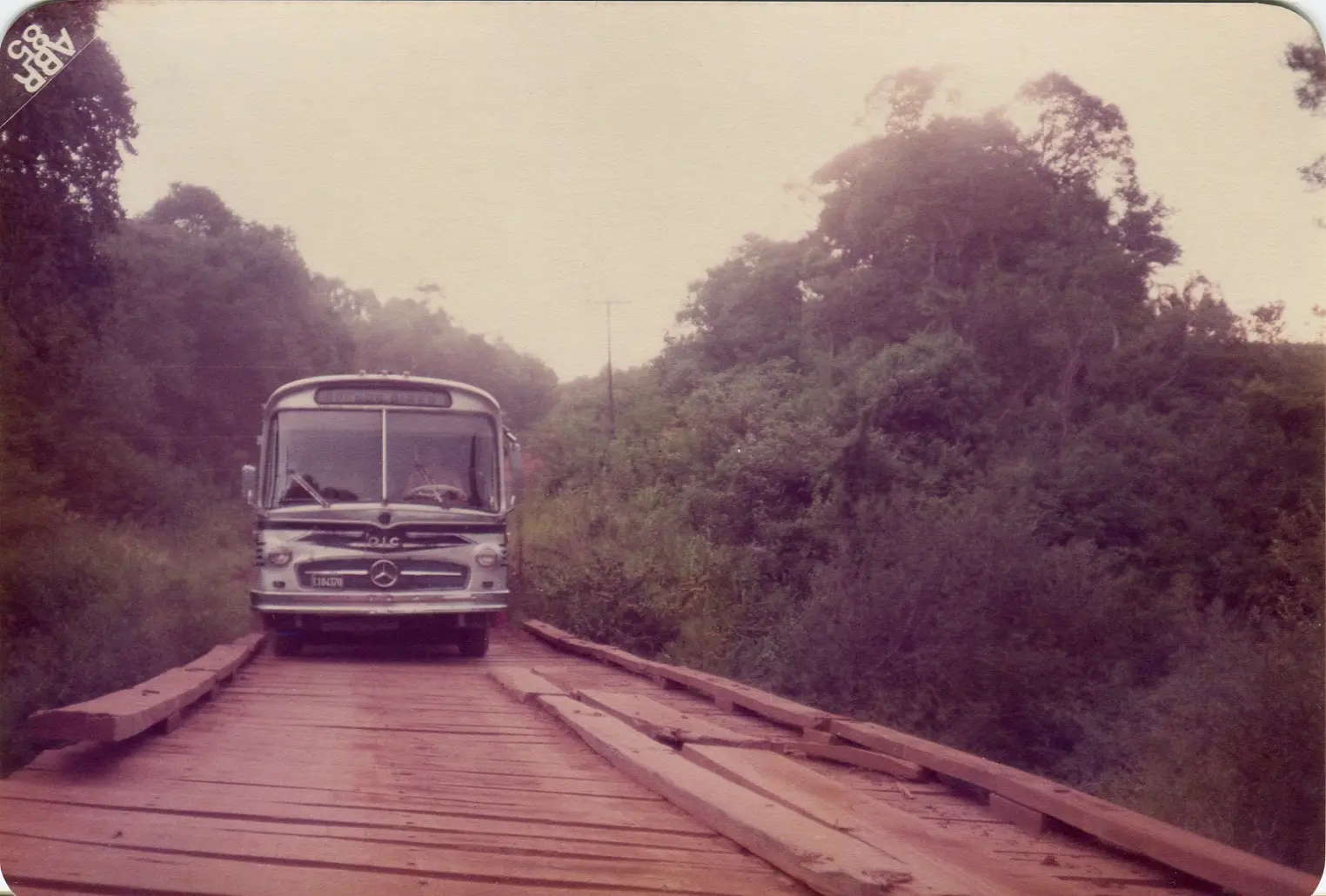 Un micro Mercedes-Benz de frente, detenido sobre un puente hecho de tablones de madera sueltos teñidos de tierra colorada, con vegetación subtropical a los dos lados