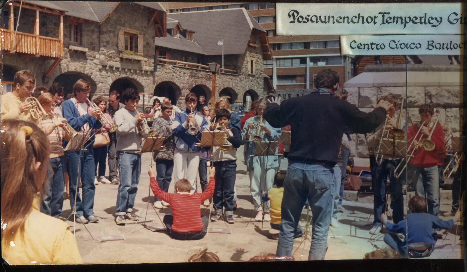 Foto panorámica del coro tocando de pie al aire libre frente a los edificios de piedra y madera del Centro Cívico de Bariloche, con chicos sentados en el suelo en primer plano