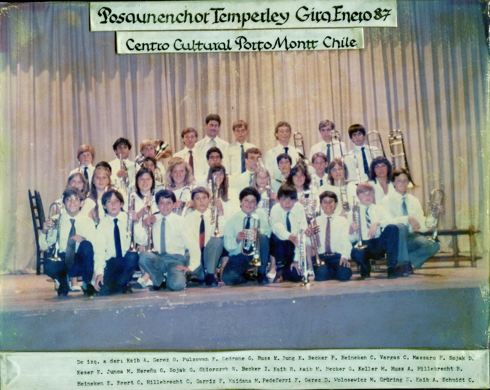 Foto grupal enmarcada de unos treinta y cinco músicos de camisa blanca y corbata sobre un escenario con cortinado, con carteles en letra gótica arriba y la nómina completa mecanografiada abajo