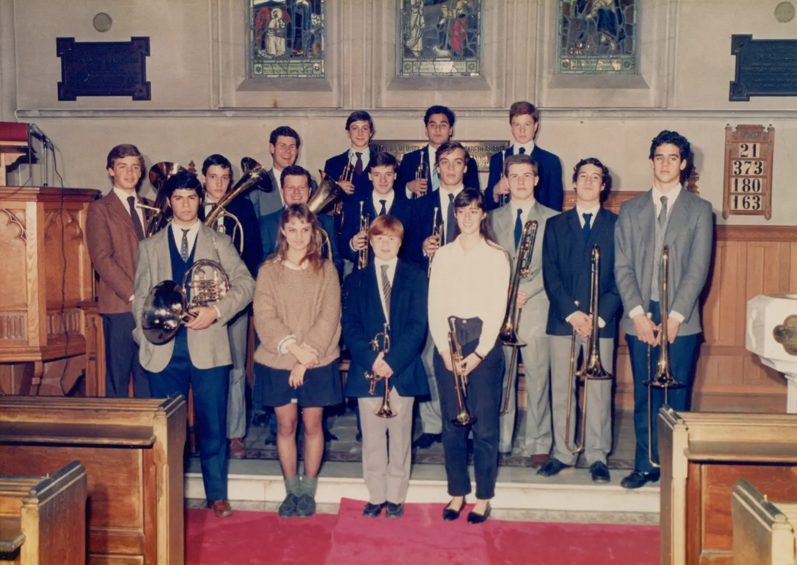 Dieciocho músicos posando con sus instrumentos en una iglesia con vitrales y placas conmemorativas en inglés