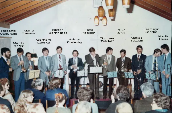 Doce músicos en fila frente al público en un salón de techo a dos aguas, foto rotulada 1980 con los nombres