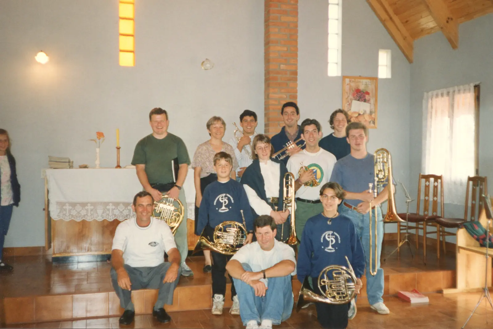 Trece músicos posando en una capilla modesta de techo de madera y pilar de ladrillo, con cornos, trompetas y trombones; dos de ellos llevan buzos azules con el escudo del coro