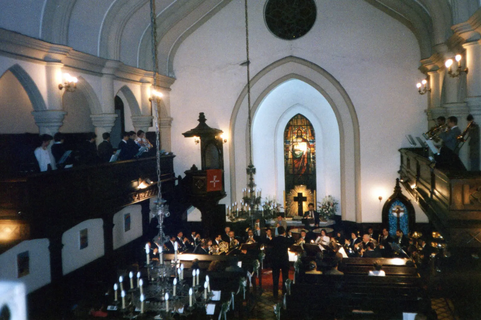 Vista desde una galería de una iglesia neogótica: el conjunto de bronces tocando en el piso con el director de pie en el pasillo, y otros grupos de músicos repartidos en las dos galerías laterales