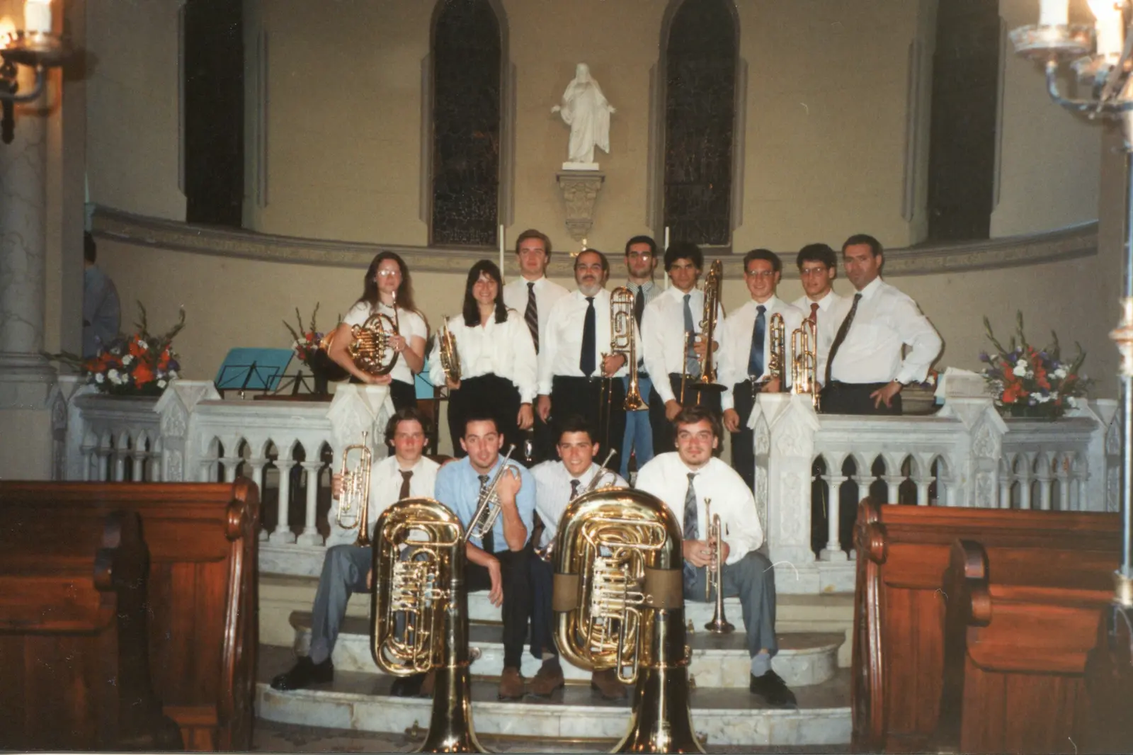 Catorce músicos de camisa blanca y corbata posando con sus instrumentos en el altar de una iglesia, con dos tubas grandes en primer plano