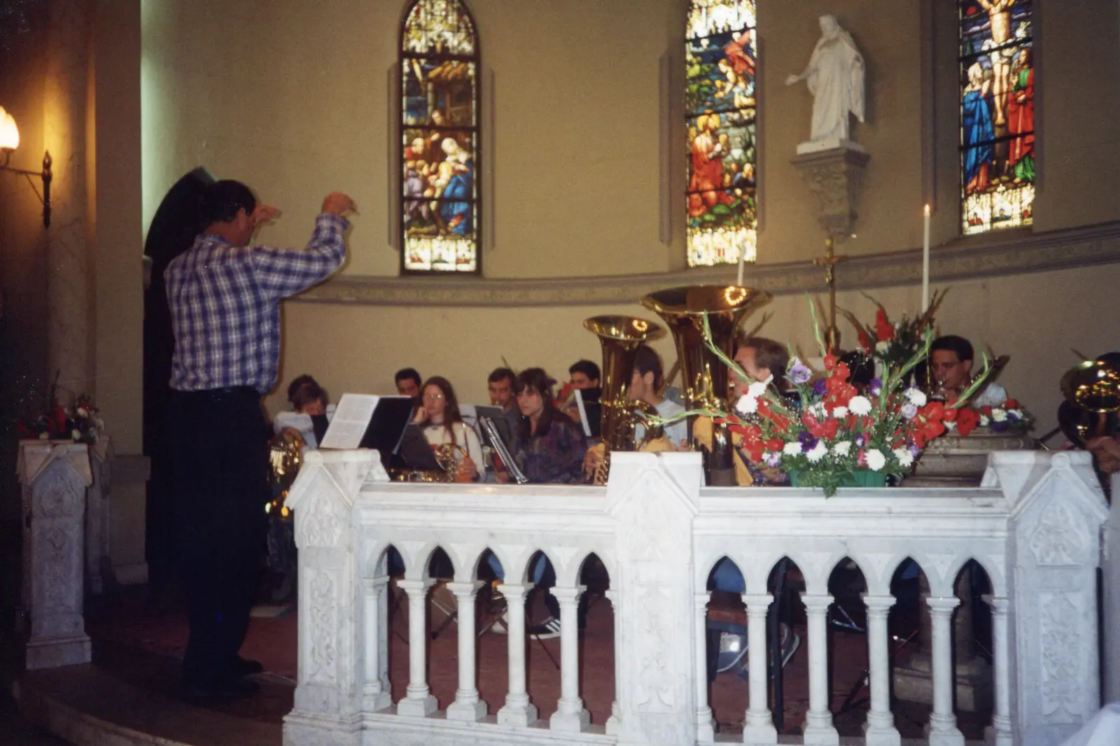 El coro tocando dentro de una iglesia con vitrales y altar de mármol, con el director de espaldas en camisa a cuadros