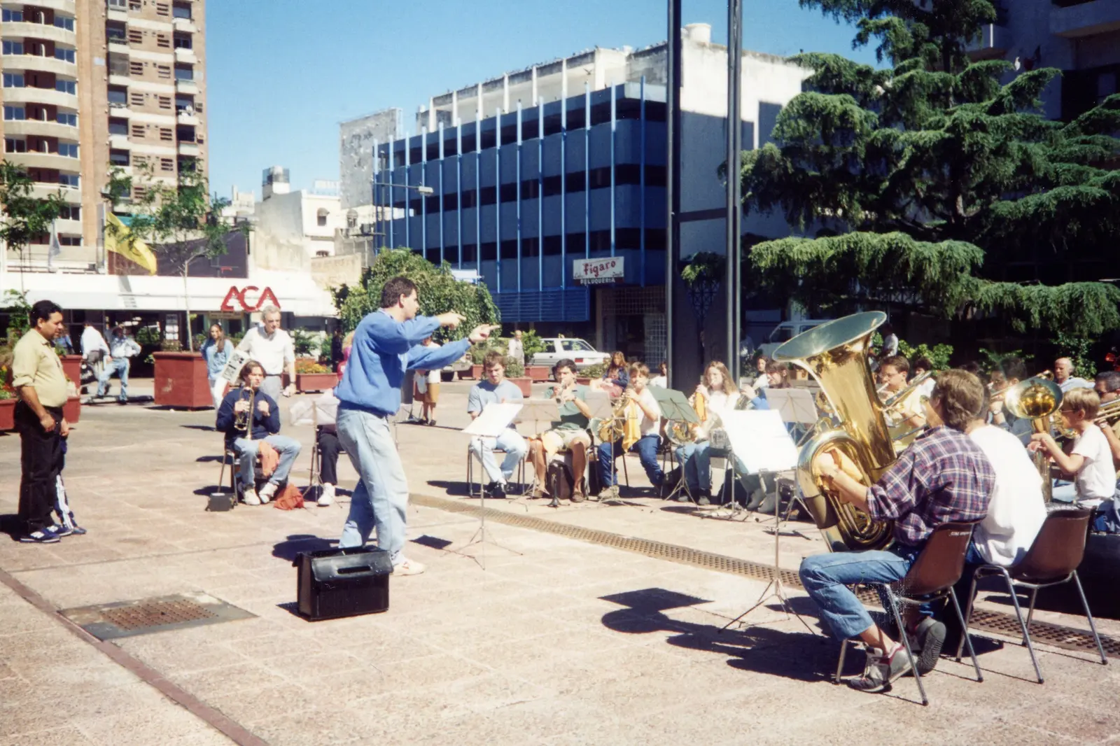 El coro tocando sentado al aire libre en una plaza de Rosario, con el director de pie sobre un cajón negro y edificios de la ciudad detrás