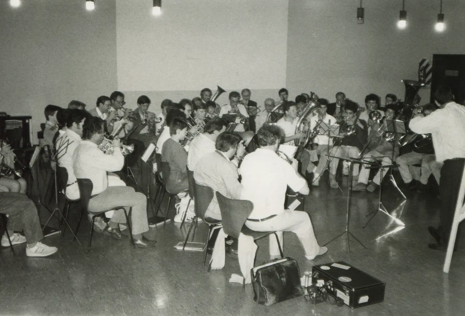 Fotografía en blanco y negro de unas cuarenta personas sentadas en filas apretadas de sillas tocando instrumentos de bronce en un salón desnudo de techo alto, con el director de pie a la derecha