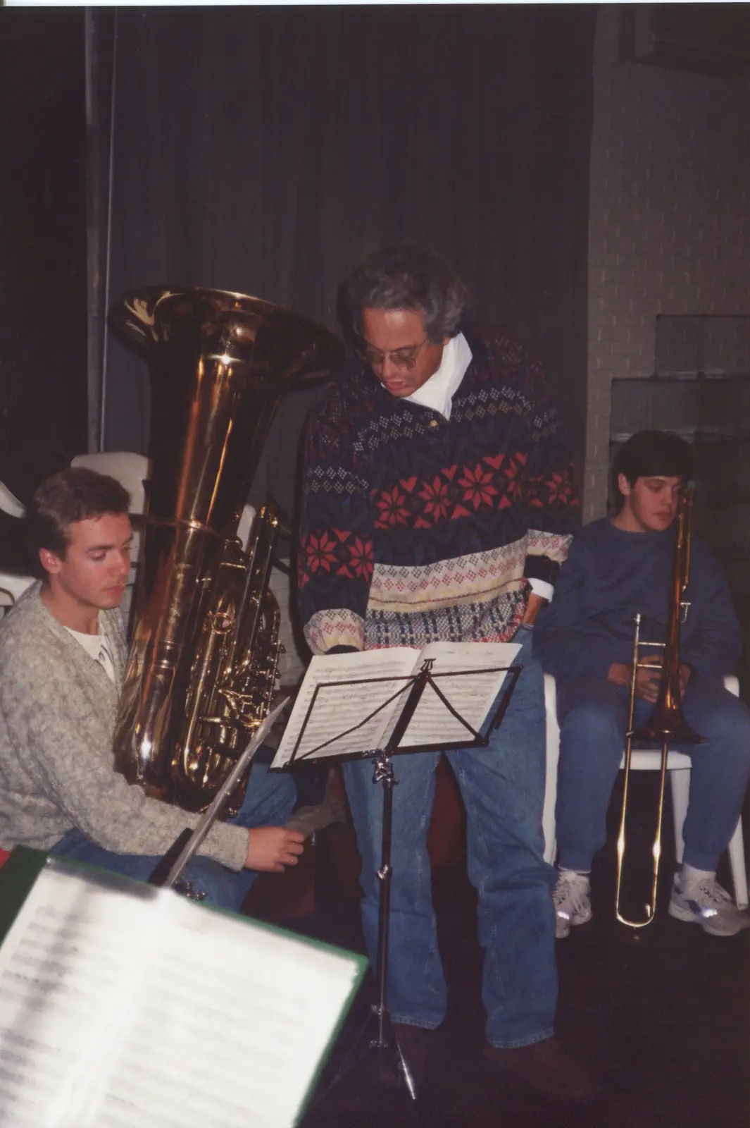 Un hombre de pelo canoso y pulóver nórdico, inclinado sobre un atril leyendo la partitura junto a un joven que sostiene una tuba