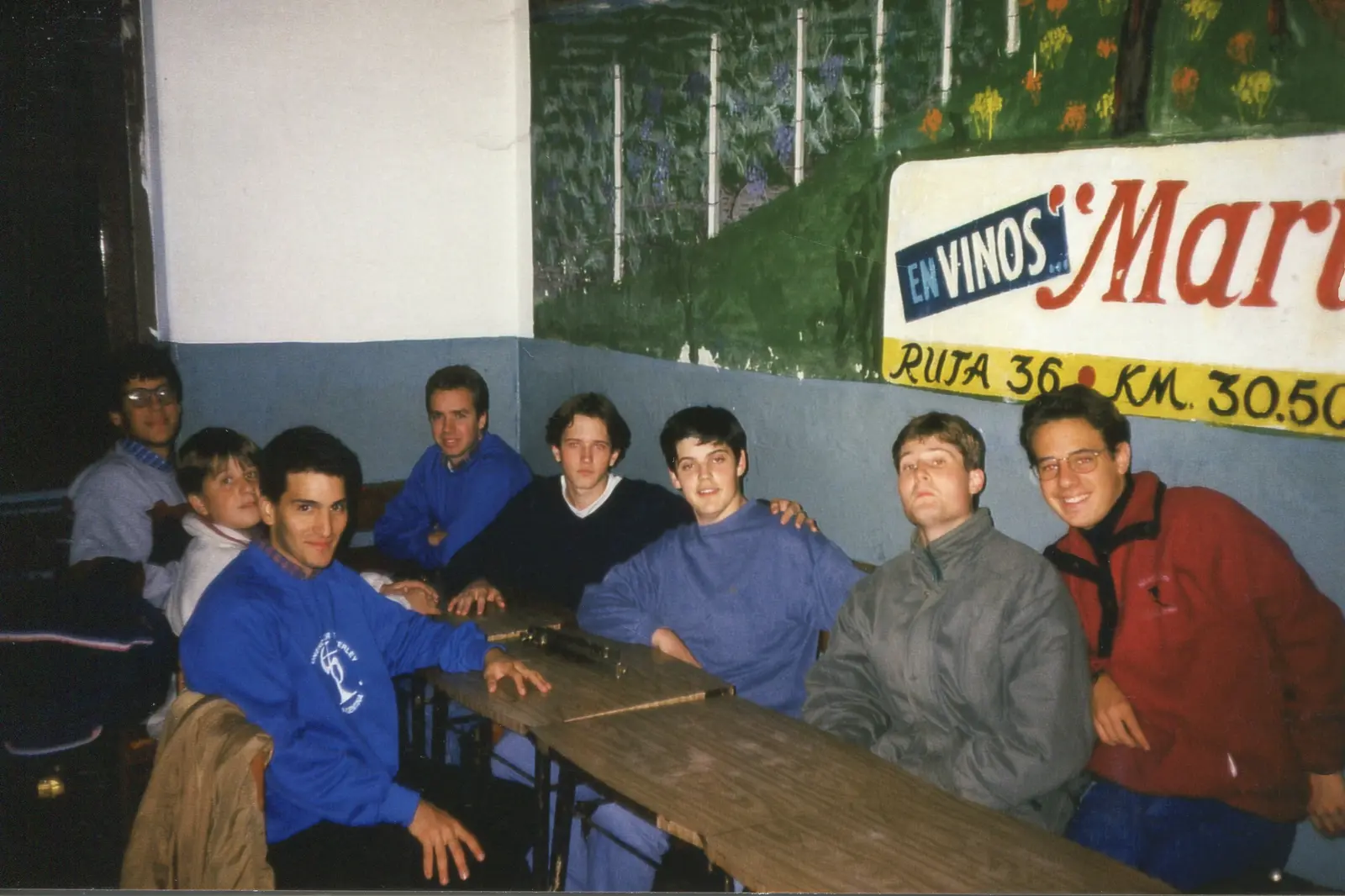 Nueve jóvenes del coro sentados a una mesa larga de un club, varios con buzos azules del Posaunenchor, con un cartel de vinos pintado en la pared