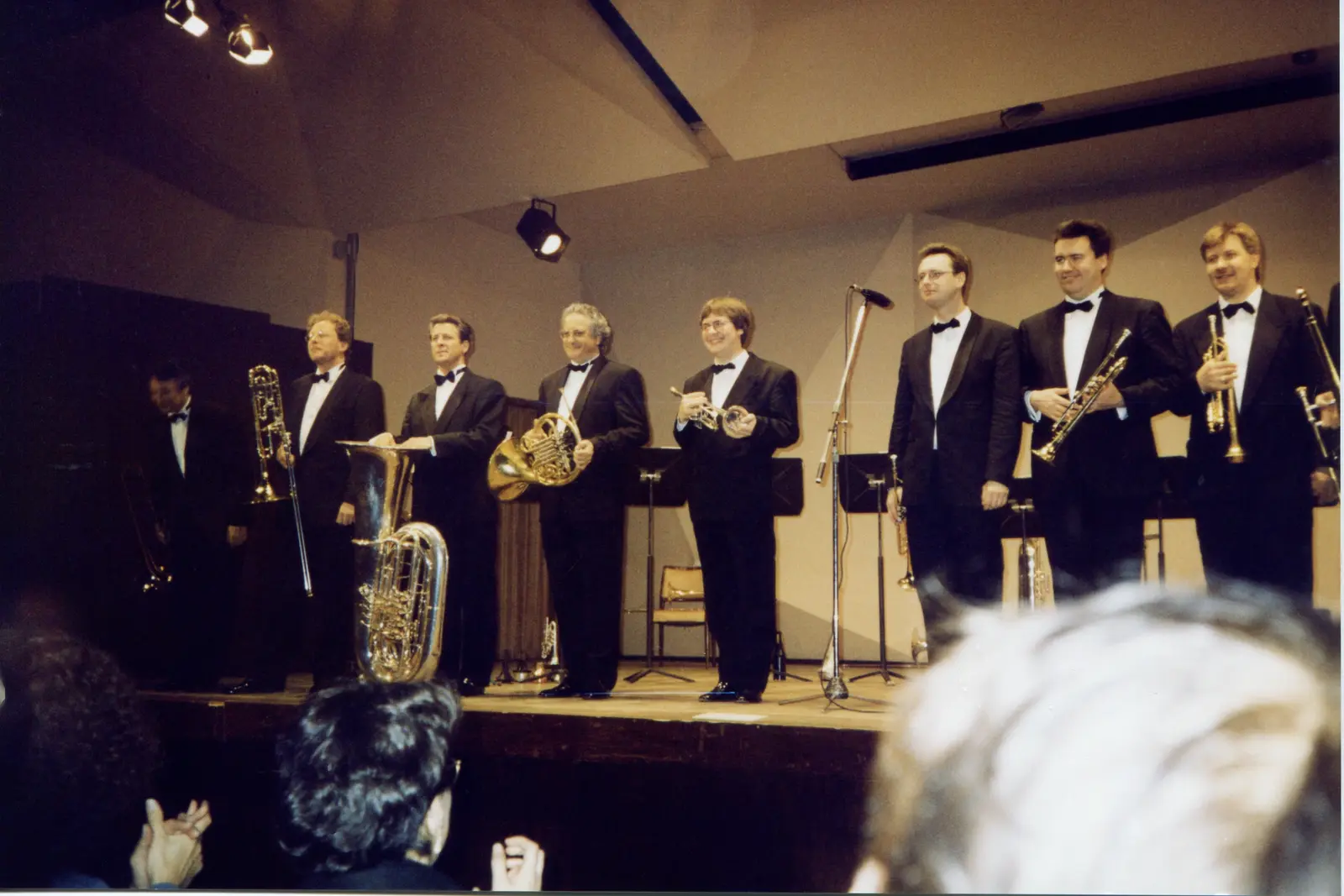 Nueve músicos de smoking saludando en el escenario con trombón, tuba, corno y trompetas, ante el público que aplaude