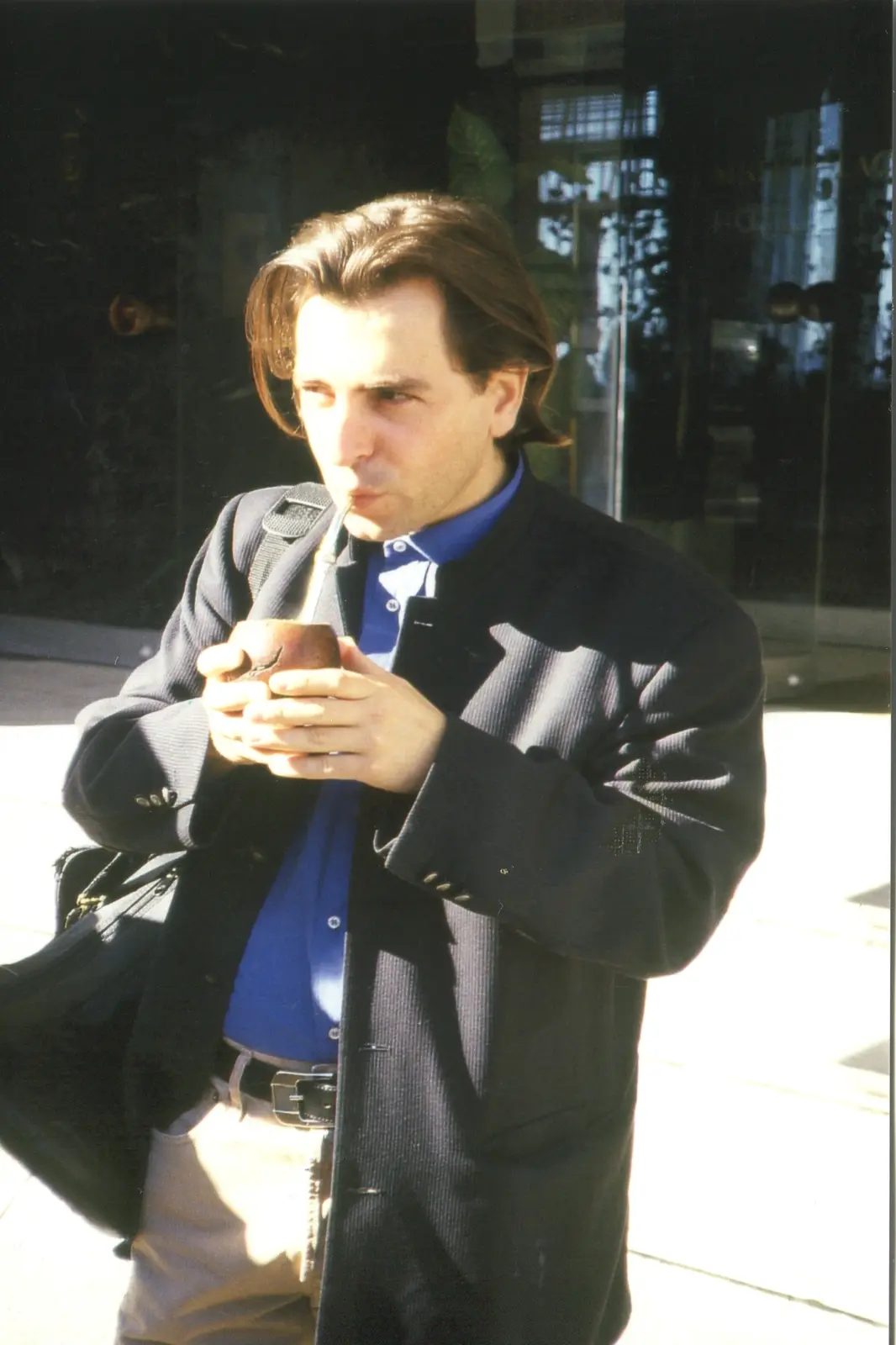 Will Sanders, de saco oscuro, tomando mate en la vereda al sol, con la bombilla en la boca y el mate entre las manos