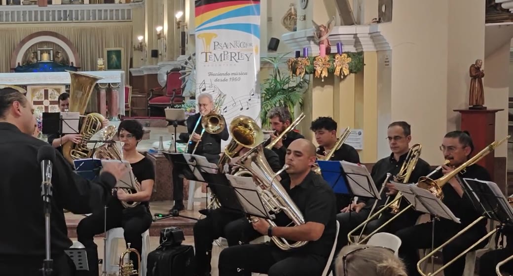 Die tiefe Blechregisterreihe des Posaunenchor Temperley beim Spiel in einer Kirche: Tuben, Euphonien und Posaunen sitzend vor den Notenpulten, im Hintergrund das Banner des Chores