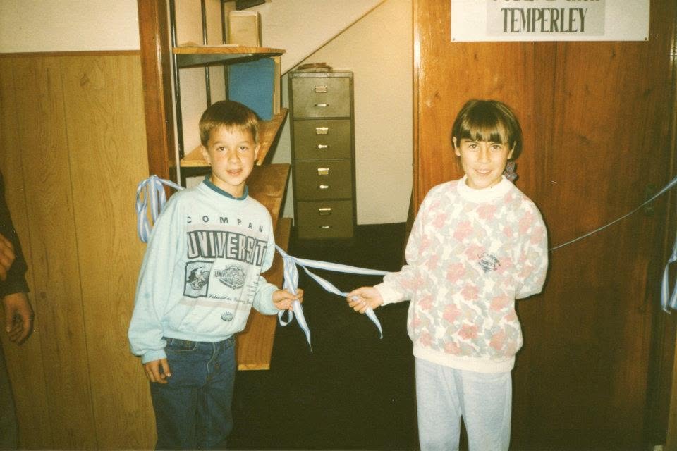 Foto en color de los años ochenta o noventa: dos chicos de unos diez años, un varón de buzo celeste y una nena de buzo con flores, sostienen cada uno una punta de una cinta celeste y blanca que acaban de cortar; detrás, un pasillo de madera con un fichero metálico y un cartel donde se lee «TEMPERLEY»