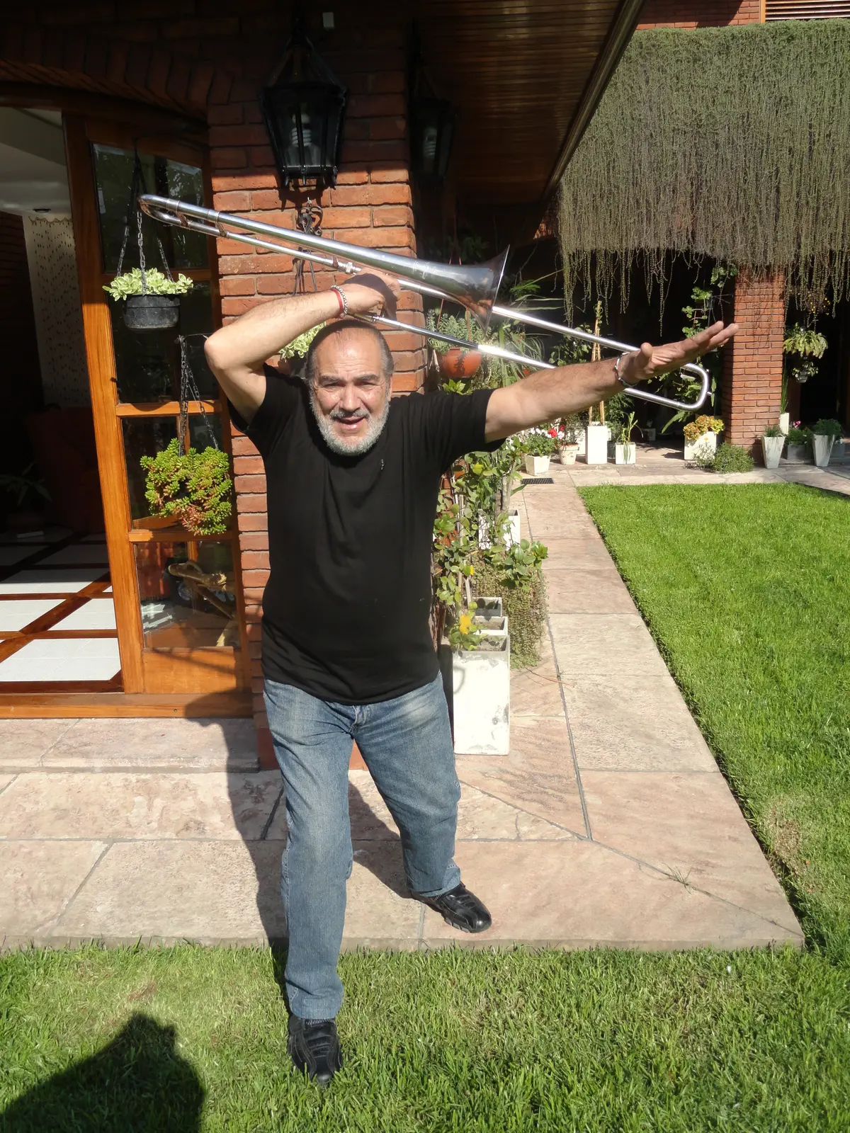 Gaspar de remera negra tocando su trombón en el jardín de una casa, con el brazo extendido en séptima posición
