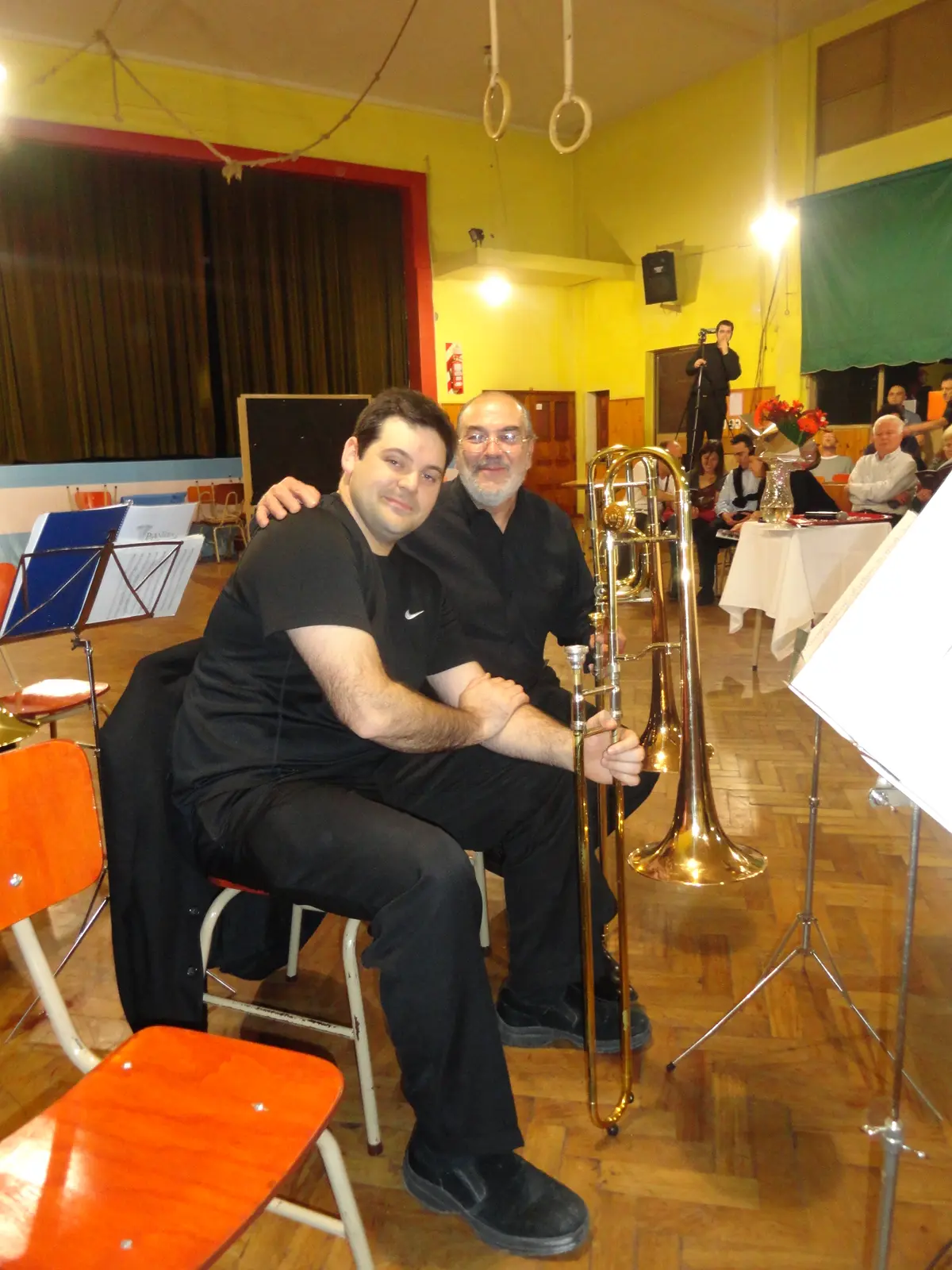 Gaspar, con barba blanca y anteojos, sentado junto a un músico joven que lo abraza, ambos de negro y con un trombón