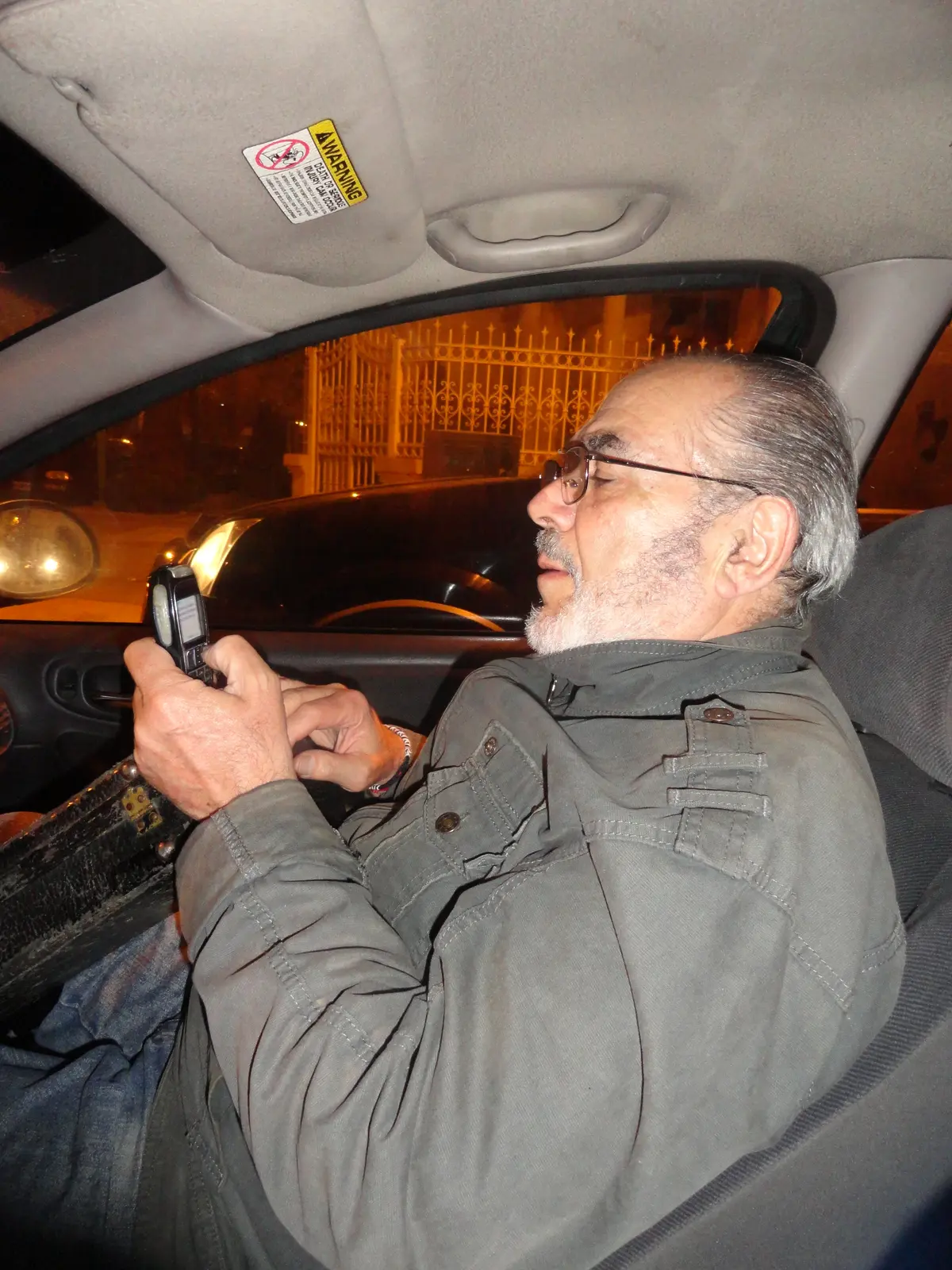 Gaspar de noche, con anteojos apoyados en la punta de la nariz, mirando la pantalla de un celular junto a un auto