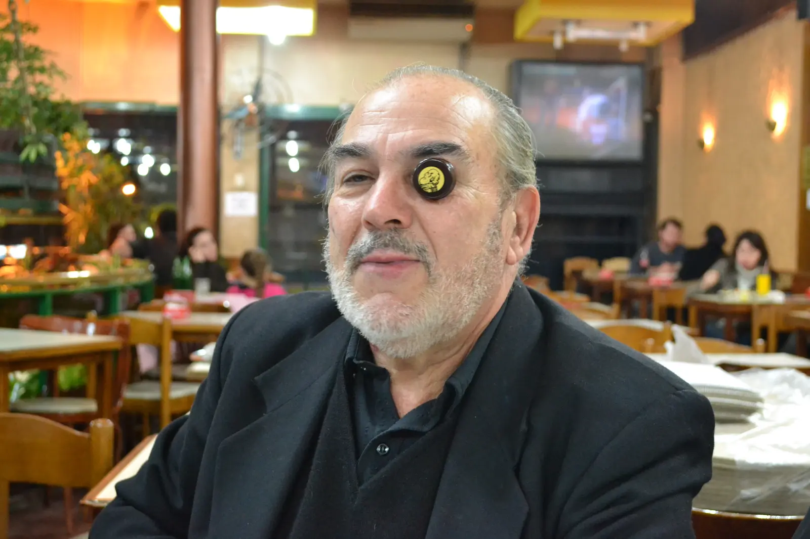 Gaspar sentado en una parrilla, de saco negro, con una boquilla de trombón apoyada en el ojo como si fuera un monóculo, mirando a cámara con cara seria