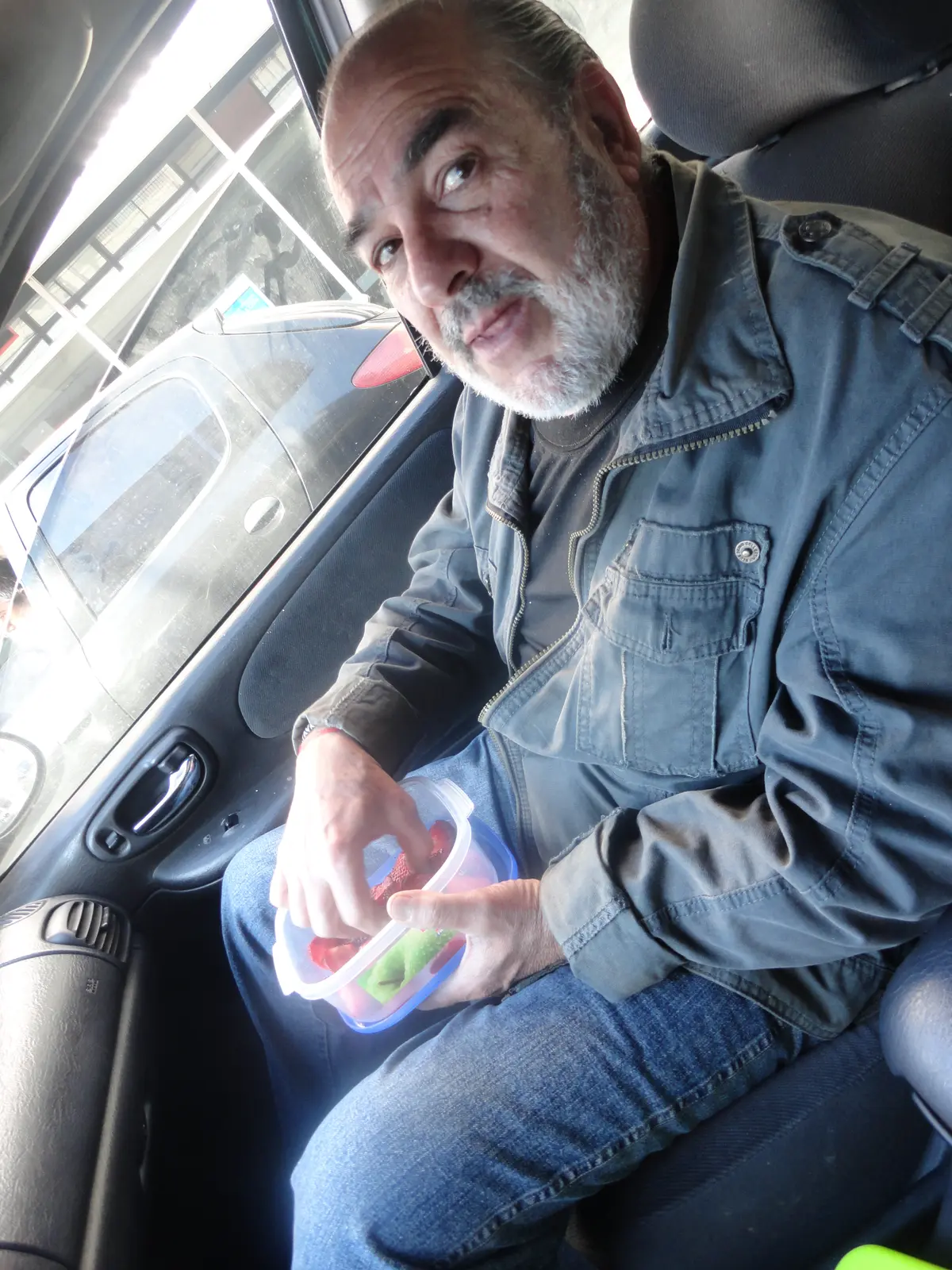 Gaspar en el asiento del acompañante de un auto, sosteniendo un tupper con fruta cortada y mirando a cámara