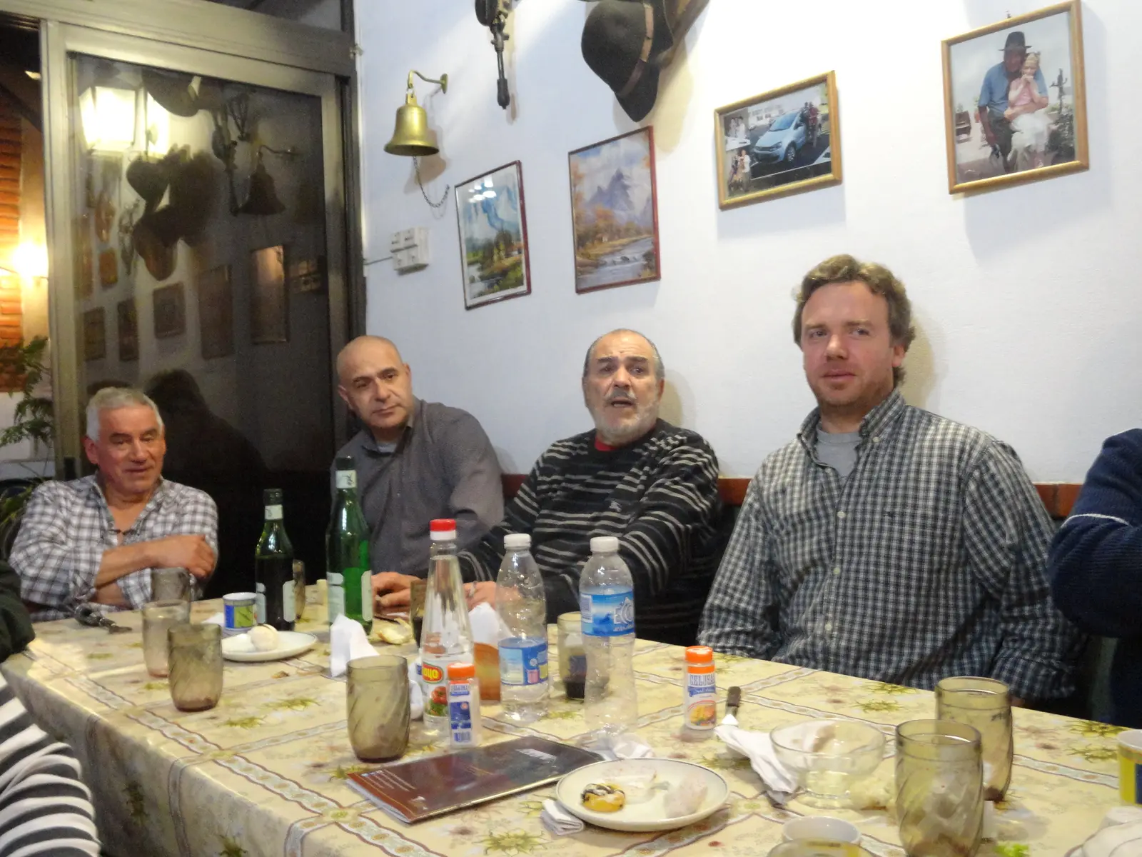 Cuatro hombres sentados a una mesa larga de restaurante con mantel floreado, botellas y vasos, mirando a cámara