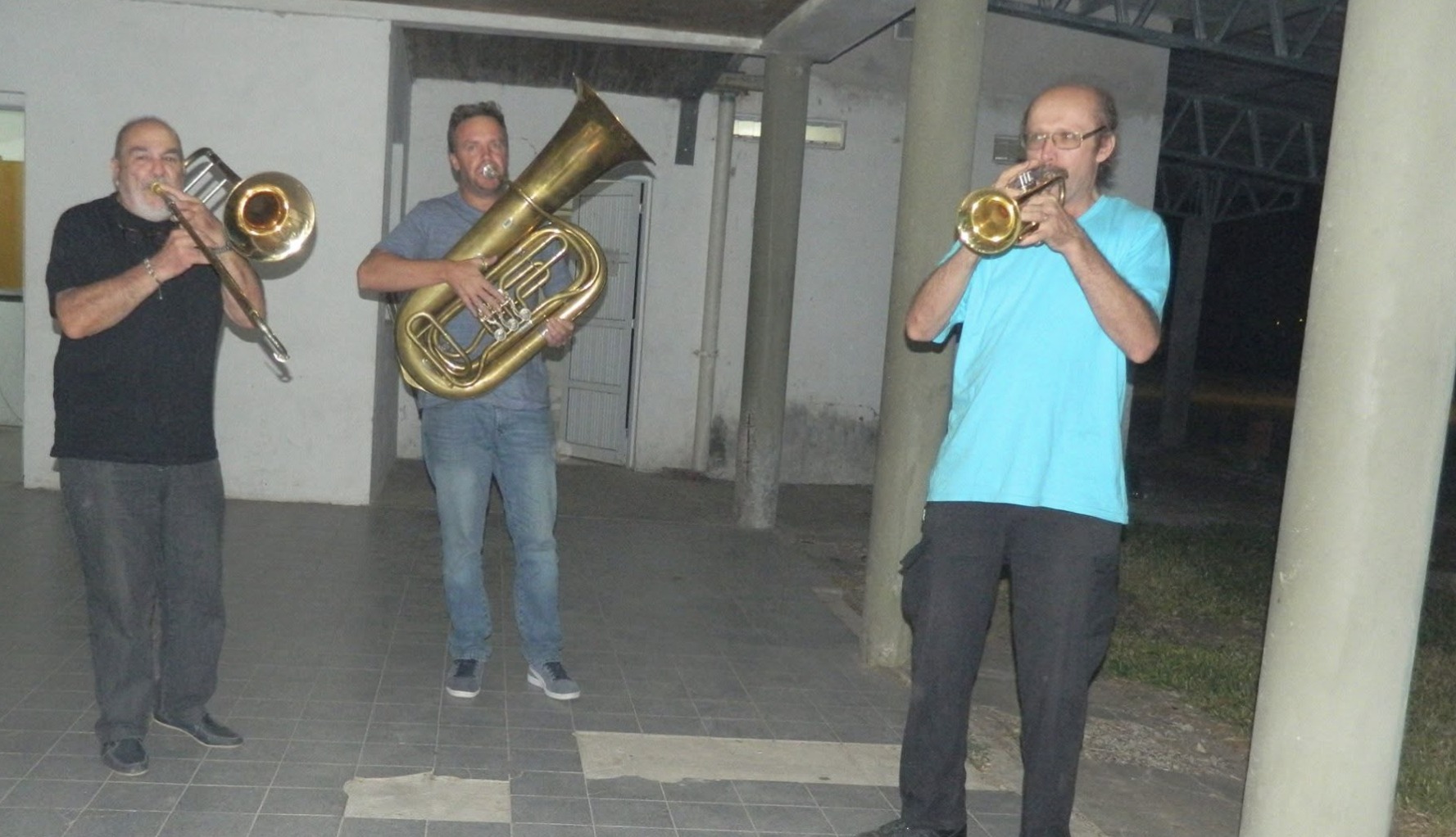 Tres músicos tocando de noche bajo una galería de baldosas: a la izquierda un hombre canoso de barba con trombón, en el medio otro de jean y remera clara con una tuba, y a la derecha un tercero de remera celeste y anteojos con una trompeta