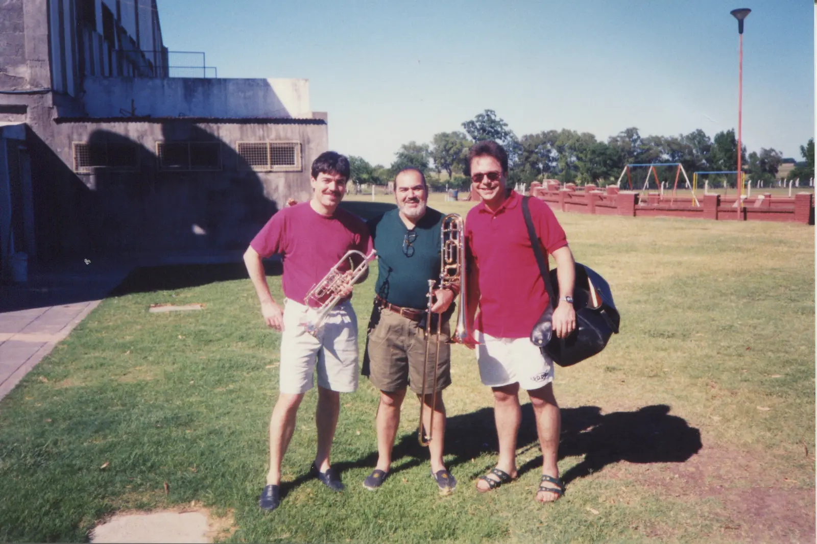 Gaspar con su trombón entre dos músicos de remera roja, uno con trompeta y otro con un bolso, en el césped de un predio