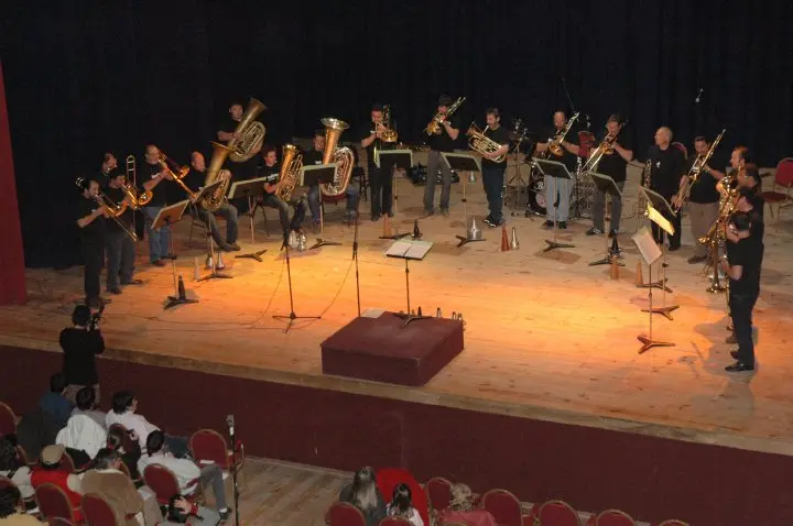 Vista desde el palco de un teatro: un ensamble de veinte músicos de bronces de negro sobre el escenario, con público en las butacas