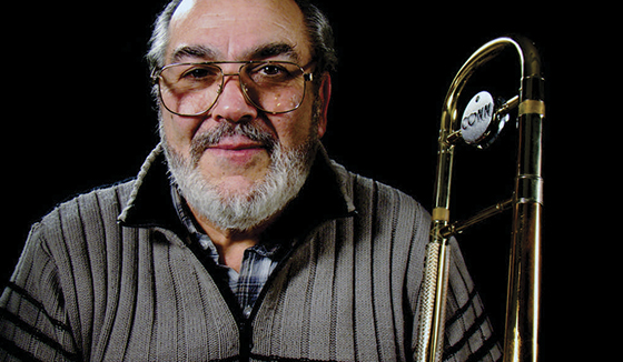 Retrato de estudio de Gaspar Licciardone con anteojos y barba blanca, junto a un trombón Conn, sobre fondo negro