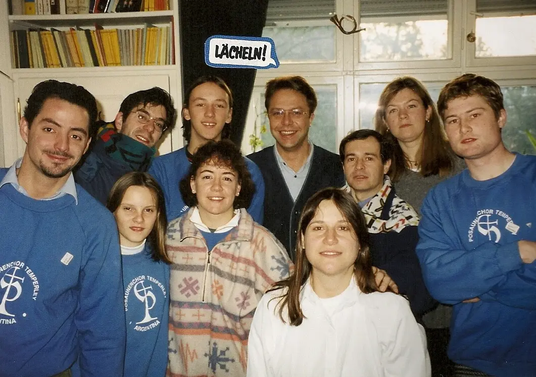 Grupo de jóvenes con buzos azules del Posaunenchor Temperley Argentina en el living de una casa, con un globo de diálogo pegado que dice Lächeln!