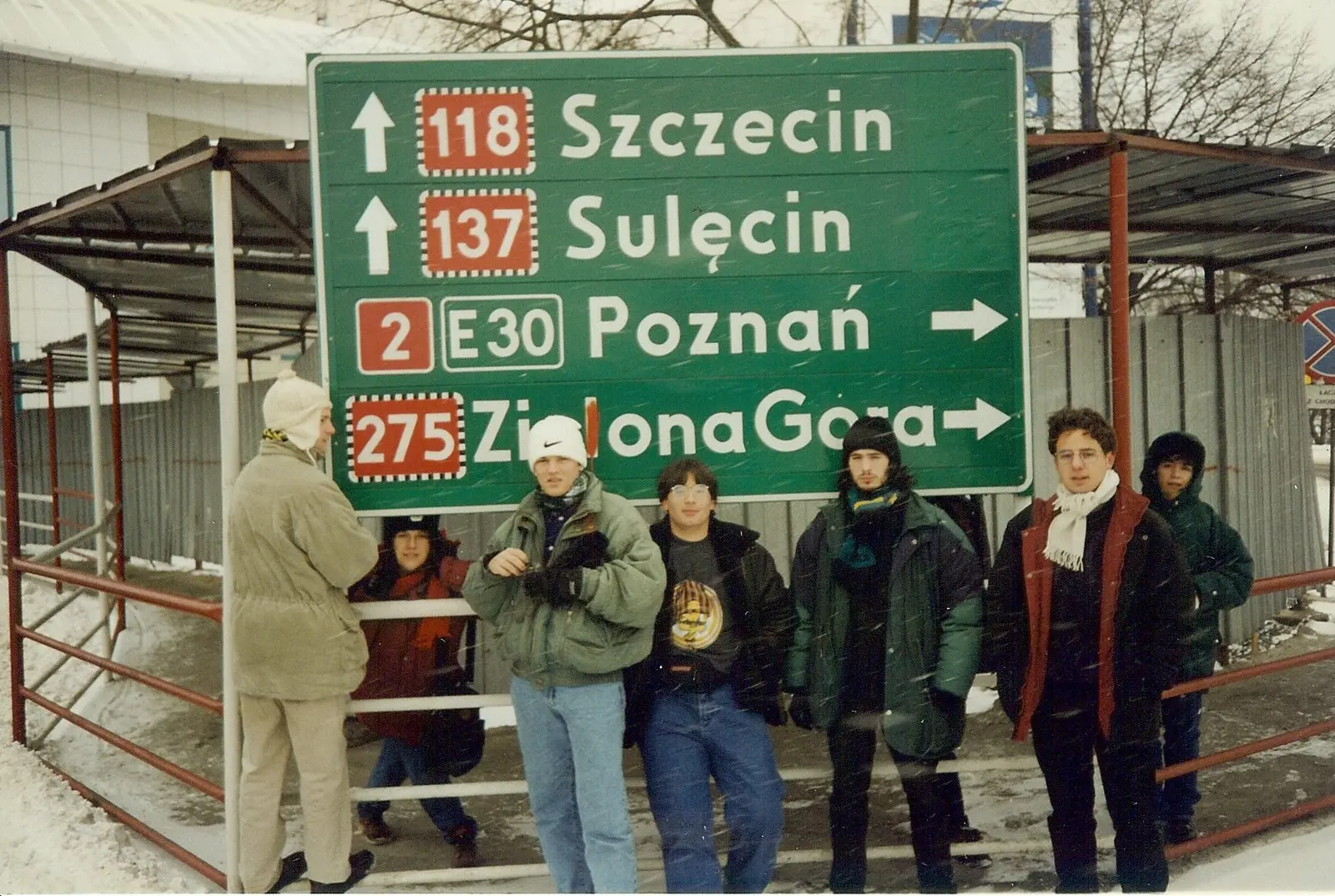 Siete jóvenes abrigados posando bajo un gran cartel vial verde en polaco que indica Szczecin 118, Sulęcin 137, Poznań por la ruta 2 E30 y Zielona Góra 275, con nieve en el suelo y cayendo