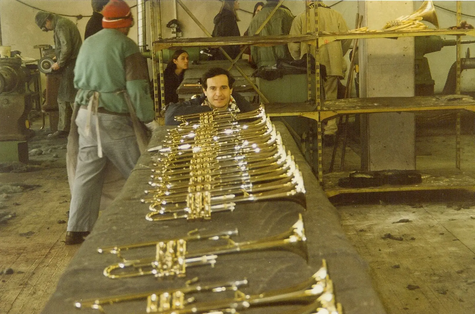 Interior de una fábrica de instrumentos de bronce: una mesa larga con veinte trompetas terminadas alineadas y brillando, operarios con delantal trabajando al fondo y un integrante del coro agachado al final de la mesa