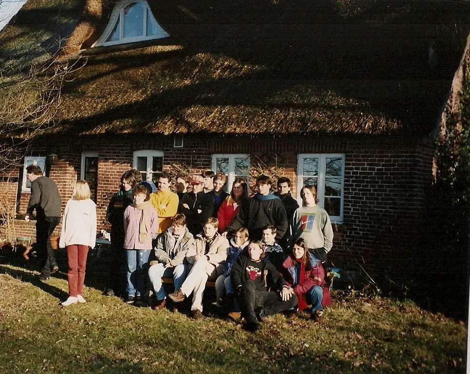 La delegación posando al sol frente a una casa de ladrillo con techo de paja, en invierno