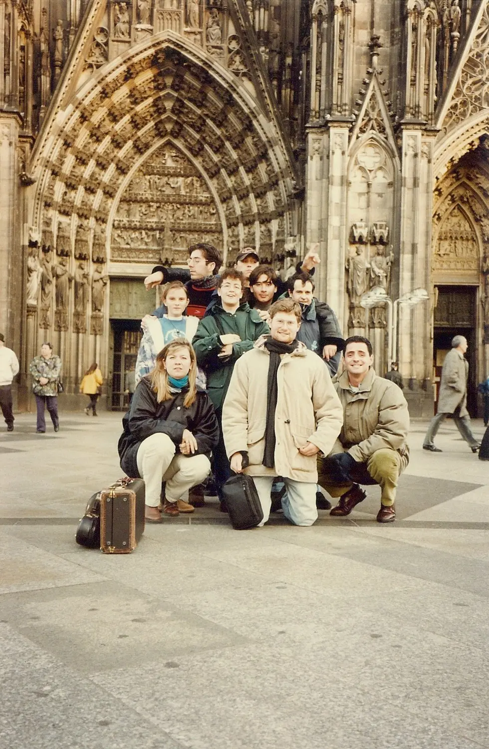 Nueve integrantes posando en cuclillas y de pie ante el pórtico gótico de la catedral de Colonia, con una valija y un estuche de instrumento en el piso