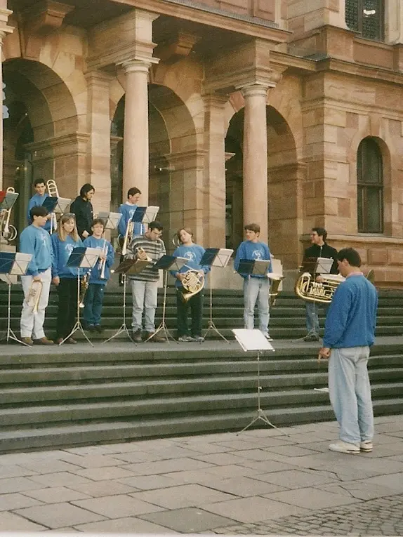 Doce músicos con buzos azules tocando de pie sobre la escalinata de un edificio de arenisca con arcadas, con atriles, y el director de espaldas dirigiendo desde la vereda