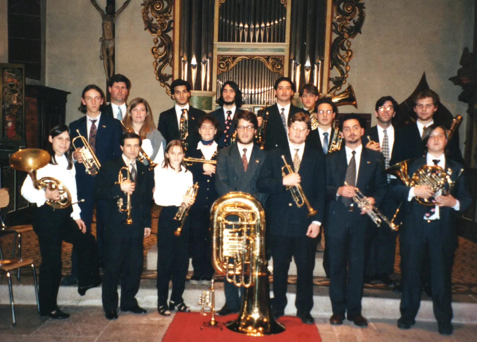 Los veinte músicos de la delegación posando de traje con sus instrumentos delante de un órgano barroco dorado, con un crucifijo de madera a la izquierda y una tuba apoyada sobre la alfombra roja