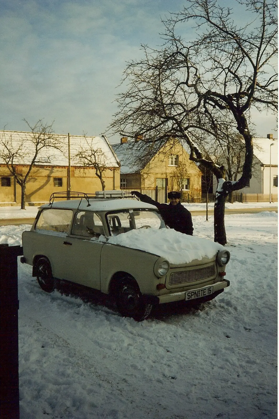 Un Trabant rural blanco con parrilla en el techo, hundido en la nieve en la calle de un pueblo de casas revocadas, con alguien de gorro de piel saludando detrás del auto