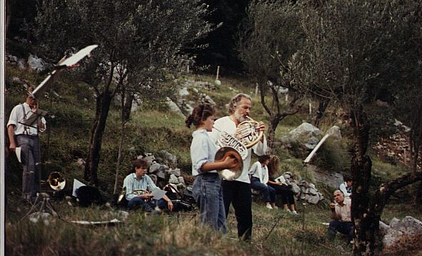 Fotografía en color de una clase de corno al aire libre en una ladera con olivos: un hombre de pelo y barba canosos toca el corno junto a una joven que sostiene otro corno, y varios músicos sentados con atriles