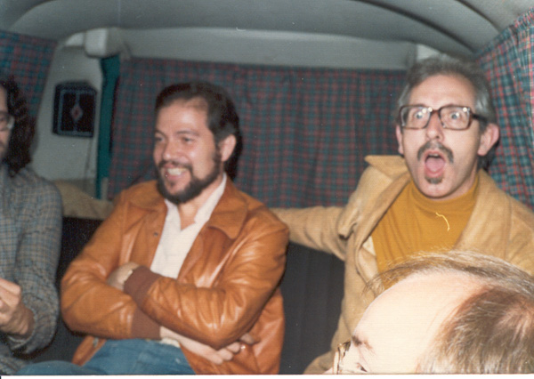 Fotografía en color, tomada dentro de una combi con cortinas escocesas: un hombre de barba y campera de cuero marrón, con los brazos cruzados, junto a otro de anteojos y bigote con suéter mostaza que gesticula