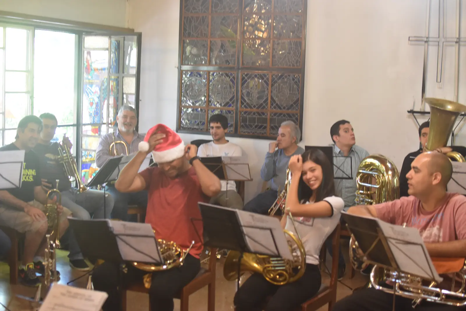 Músicos de bronce sentados con atriles en un salón; uno se acomoda un gorro de Papá Noel