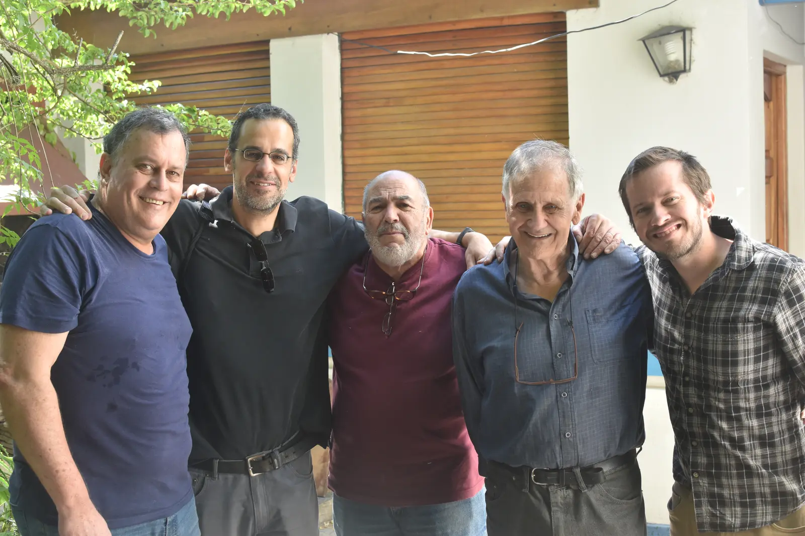 Cinco hombres abrazados posando en un patio, delante de una casa con persianas de madera y un farol