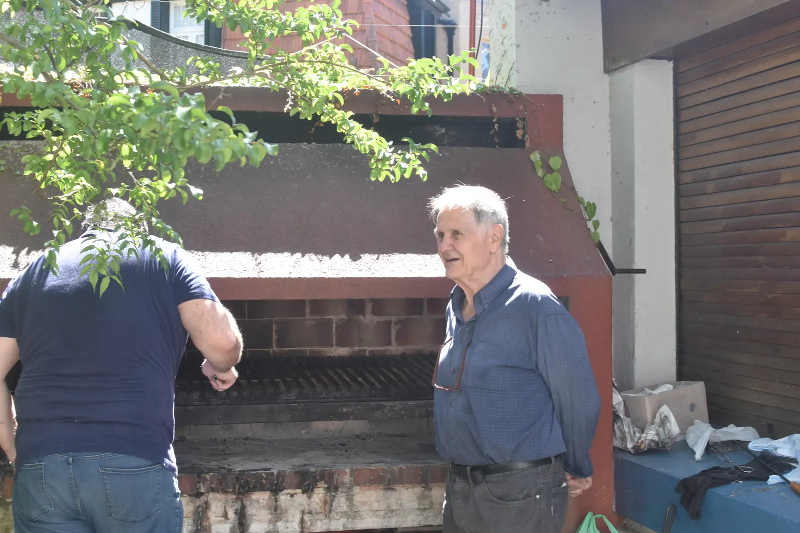 Dos hombres junto a una parrilla de ladrillo bajo una enredadera